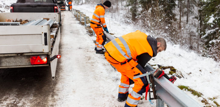 Arbetare i orange varselkläder reparerar ett vägräcke vid en snötäckt väg.