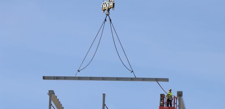 En balk lyfts av en lyftkran. Nedanför, men inte under, står vad som ser ut att vara en man i bygghjälm och arbetskläder.