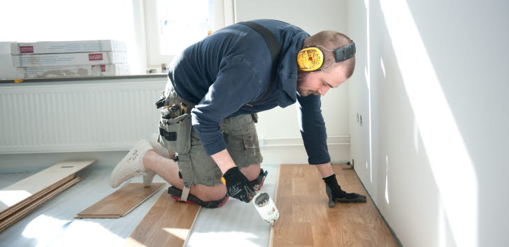 En person installerar trägolv inomhus, och bär hörselskydd och en verktygsbälte.