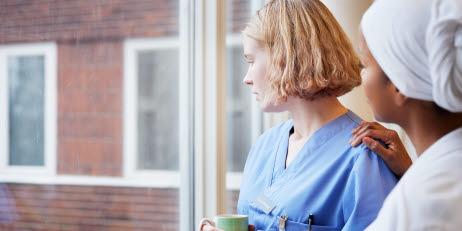 Två sjuksköterskor tittar ut genom ett fönster. Den ena personen stöttar den andra genom att lägga en hand på axeln.