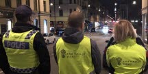 En polis och två arbetsmiljöinspektörer i gula varselkläder, fotograferade bakifrån på en gata