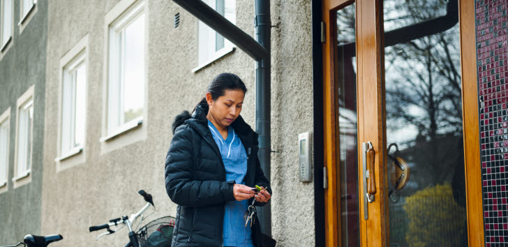 En kvinna som arbetar inom hemtjänsten står utanför en port till ett flerfamiljshus. Hon håller i en nyckelklippa. Bakom henne står en cykel.