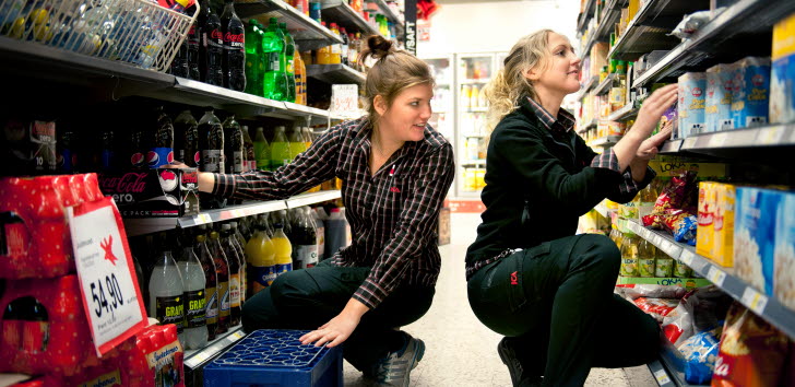 Två unga kvinnor som står på huk mellan två butikshyllor och plockar med varor