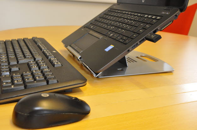 Bärbar dator på laptopställning samt tangentbord och mus liggandes på bordet.