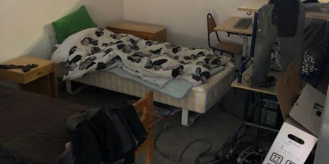 Boende på jobbet, stökigt rum med säng, skrivbord, sladdar och kartonger.