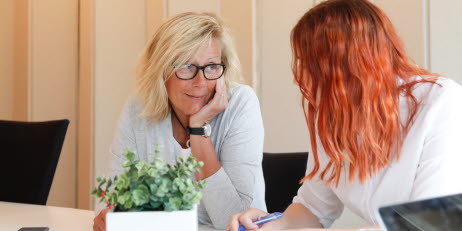 Två personer sitter vid ett bord i kontorsmiljö och samtalar. En krukväxt står på bordet.