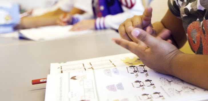 Bild på ett barn som sitter med en skolbok och räknar på fingrarna, i bakgrunden syns ett annat barn.