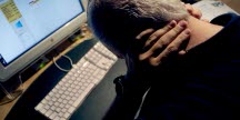En gråhårig man fotad bakifrån som sitter böjd över ett tangentbord och håller händerna i nacken.