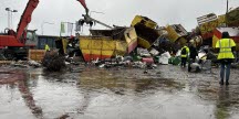 En skrotningsplats med en grävmaskin med gripklo som lyfter metallskrot. Containrar och metallavfall ligger utspridda på en våt gårdsplan. Flera personer i varselkläder står i förgrunden.