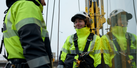 Två personer med gula skyddsjackor och personlig fallskyddsutrustning arbetar på höjd vid en byggnad.