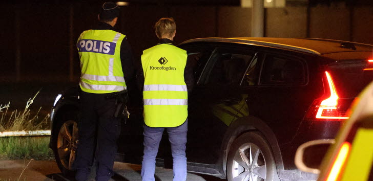 Två personer står framför en bil med ryggen vänd mot kameran. Personerna har gula västar med texten "polisen" och "kronofogden".