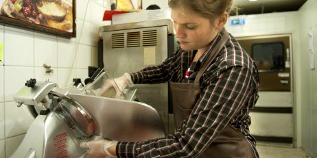 En ung person i förkläde skivar kött med en skärmaskin i en mataffär.