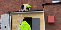 Två personer som arbetar på ett tak, mot byggnaden står en stege lutad. Nedanför syns en arbetsmiljöinspektör bakifrån.