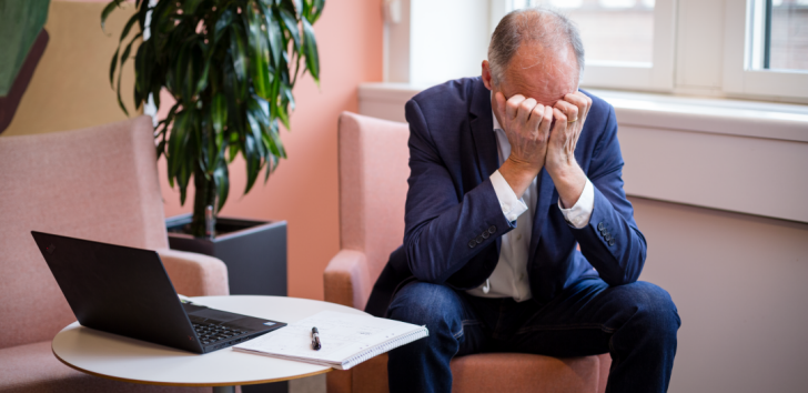 En man i kostym sitter på ett kontor och lutar sitt huvud i händerna bredvid en öppen laptop och ett anteckningsblock.