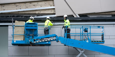 Två byggnadsarbetare står i en skylift och utför arbete på en fasad. I en höj- och sänkbar ställning bredvid står deras kollega och lutar sig mot räcket.