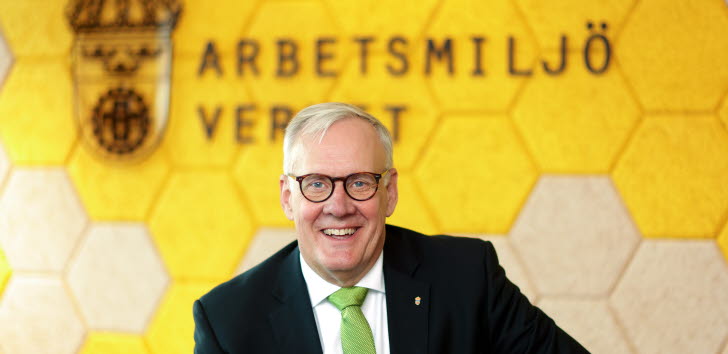 En man i kostym står lutad mot ett skrivbord och tittar in i kameralinsen. 