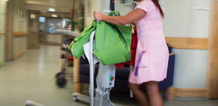 Kvinna i rosa vårduniform flyttar en lyftanordning i en sjukhuskorridor.