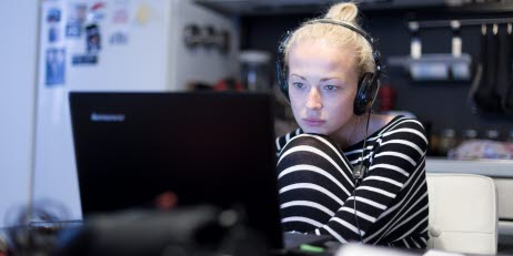 Kvinna sitter uppkrupen i pyjamas i sitt kök och arbetar med en laptop.