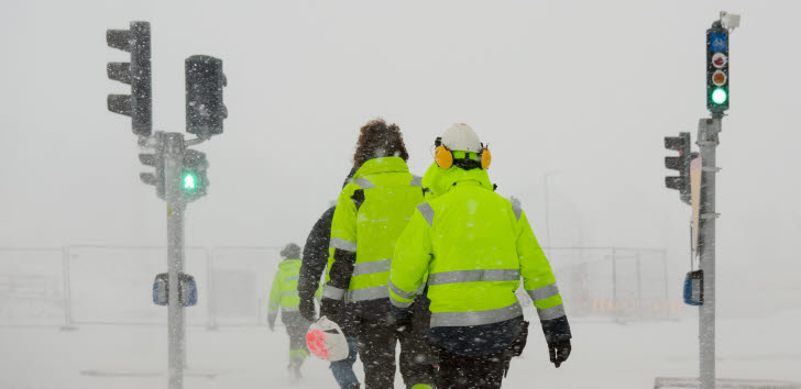 Personer i varselkläder korsar ett övergångsställe i snöyra. 