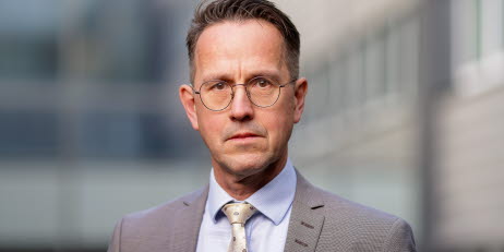 Porträtt av Arbetsmiljöverkets generaldirektör Lars Lööw i kostym och slips med glasögon, mot en suddig bakgrund av en byggnad.