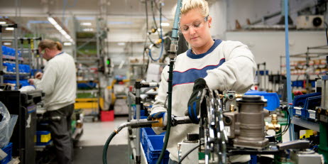 En kvinna med skyddsglasögon och skyddshandskar arbetar med en maskin och en man i bakgrunden arbetar vid en arbetsbänk.