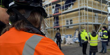 Arbetsmiljöinspektörer står med ryggen empt fotografen i för- och bakgrund på en byggarbetsplats med ställningar på en husvägg.