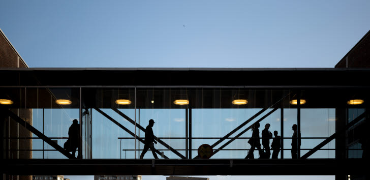 Personer går över en inglasad gångbro mot en klarblå himmel, med stadssilhuett i bakgrunden.