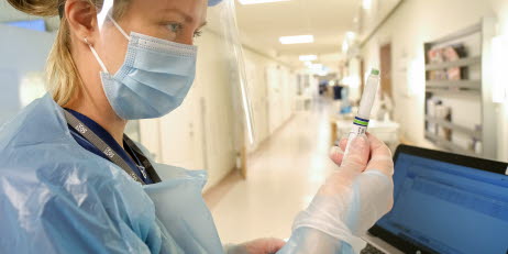 En kvinna med munskydd och visir som står i en sjukhuskorridor och håller i en kanyl