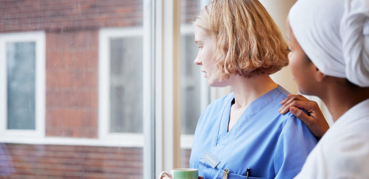 Två sjuksköterskor tittar ut genom ett fönster. Den ena personen stöttar den andra genom att lägga en hand på axeln.