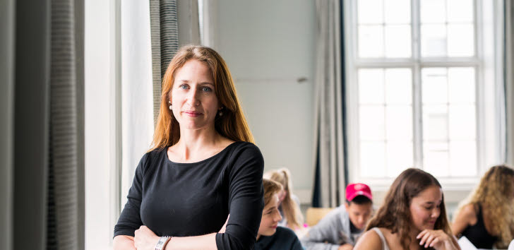 En lärare står i ett klassrum. Bakom henne sitter elever vid sina bänkar.