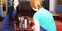 Två unga kvinnor i förkläden hanterar kaffetermosar och koppar i en kafeteria.