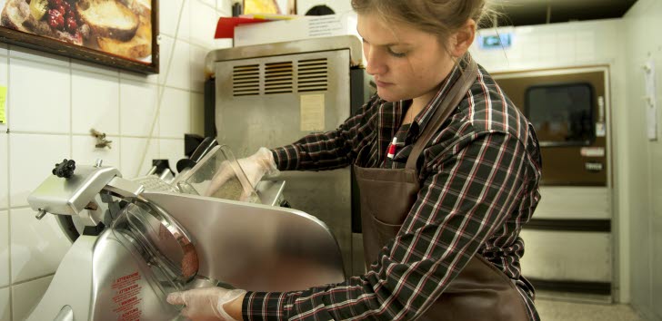 En ung person i förkläde skivar kött med en skärmaskin i en mataffär.