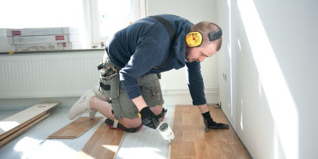 En person installerar trägolv inomhus, och bär hörselskydd och en verktygsbälte.