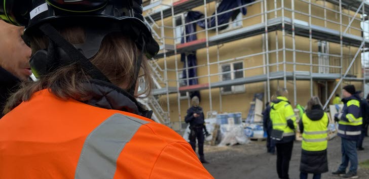 Arbetsmiljöinspektörer står med ryggen empt fotografen i för- och bakgrund på en byggarbetsplats med ställningar på en husvägg.