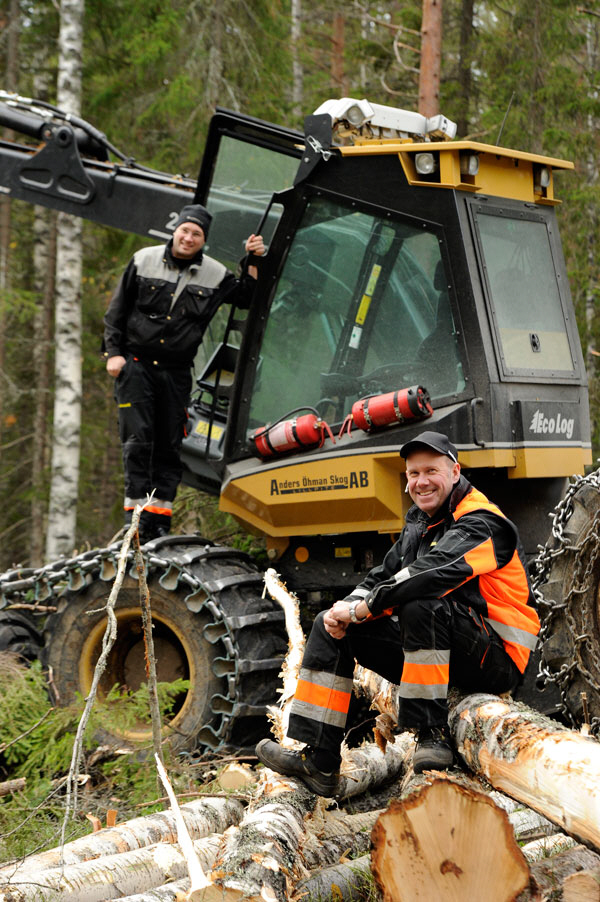 Bild på två glada skogsarbetare vid skogsmaskin bland trästockar