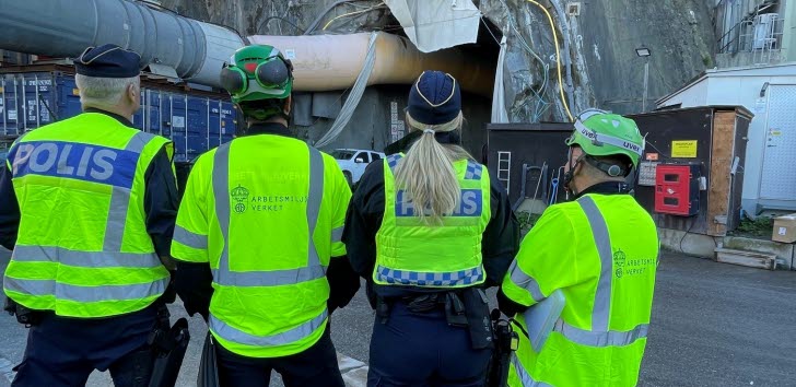 Fyra personer i reflexvästar från polisen och Arbetsmiljöverket utför en inspektion utanför en tunnel på en byggarbetsplats.