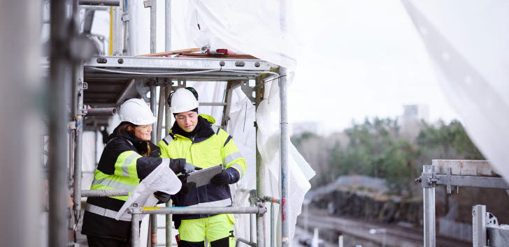 En man och en kvinna iförda bygghjälm och byggkläder står uppe i en byggställning och tittar ner i ett anteckningsblock som kvinnan pekar på.