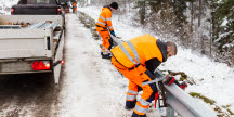 Arbetare i orange varselkläder reparerar ett vägräcke vid en snötäckt väg.
