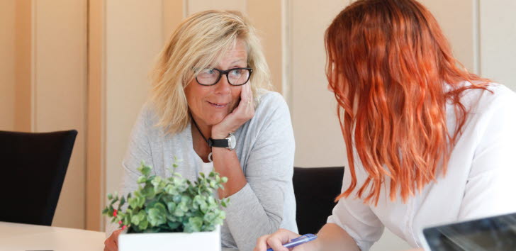 Två personer sitter vid ett bord i kontorsmiljö och samtalar. En krukväxt står på bordet.