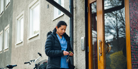 En kvinna som arbetar inom hemtjänsten står utanför en port till ett flerfamiljshus. Hon håller i en nyckelklippa. Bakom henne står en cykel.