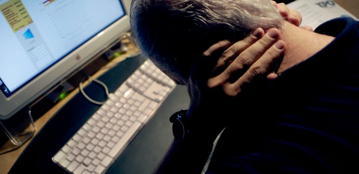 En gråhårig man fotad bakifrån som sitter böjd över ett tangentbord och håller händerna i nacken.
