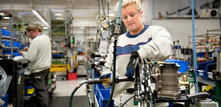 a woman with safty goggles and safty gloves working on a machine and a man in the background working at a workbench