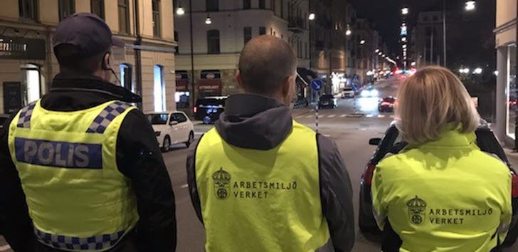 En polis och två arbetsmiljöinspektörer i gula varselkläder, fotograferade bakifrån på en gata