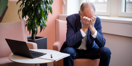 En man i kostym sitter på ett kontor och lutar sitt huvud i händerna bredvid en öppen laptop och ett anteckningsblock.